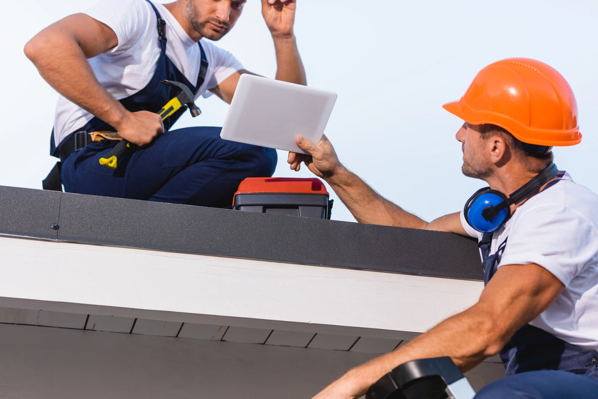 Two general contractors with hard hats working on a roof, where one is showing the other something on a tablet.