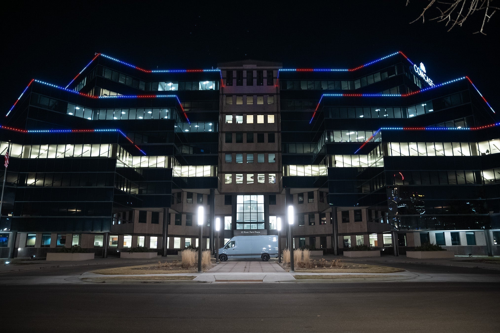 A sprawling, 9-story business building at night with rows of blue and red Christmas lights glowing along the edges of its floors, and a Comcast sign at the top.